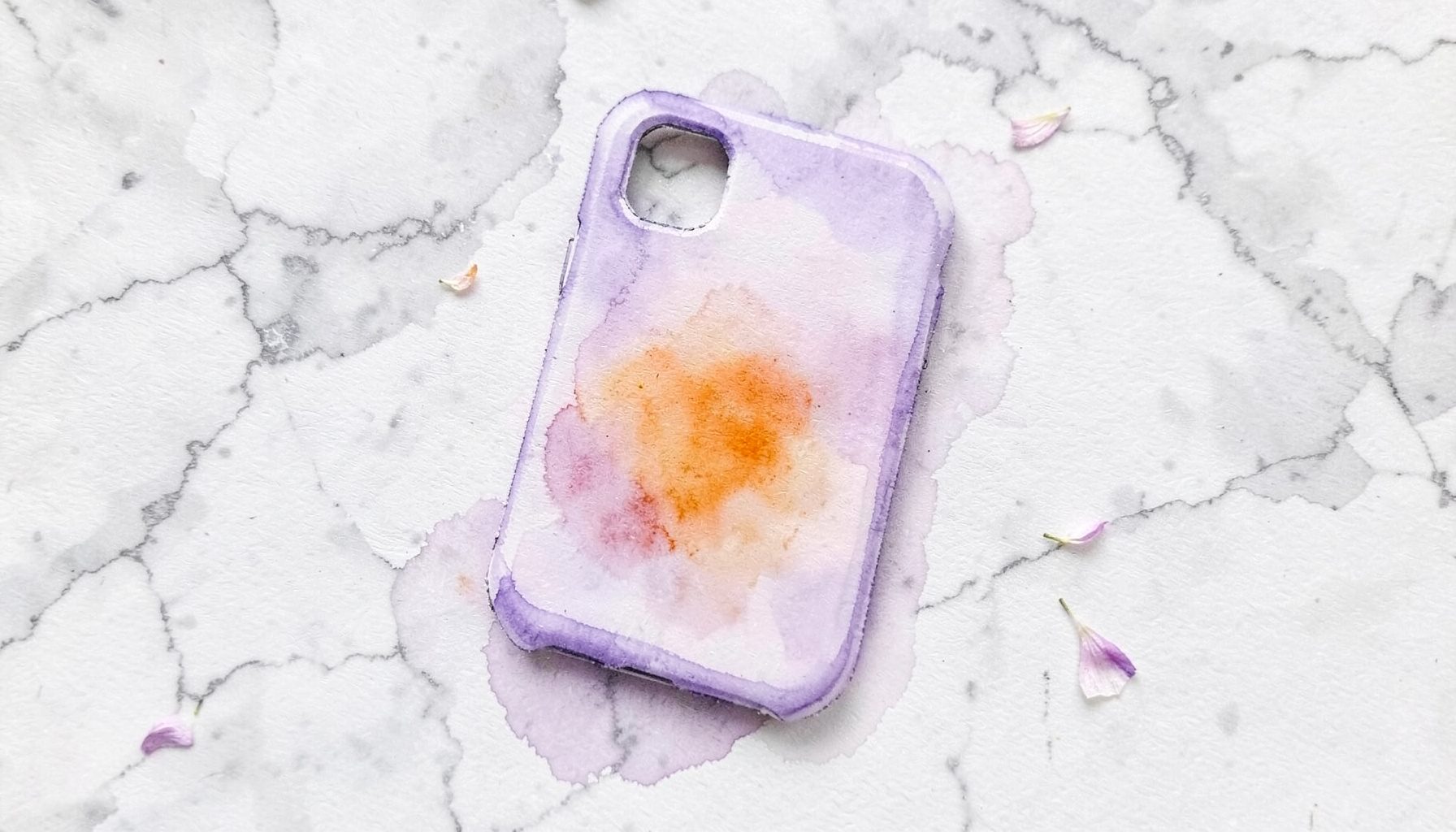 A soft watercolour painting of a personalized phone case in soft lavender tones, placed on a marble surface with a few scattered flower petals.